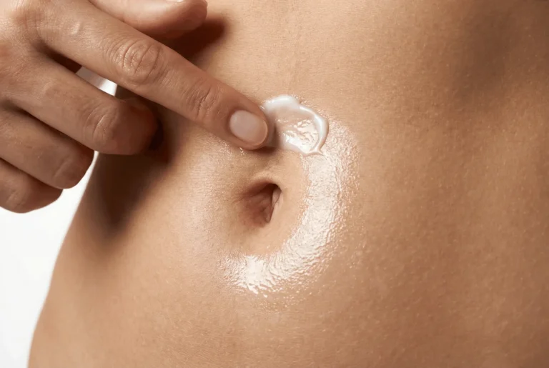 Close-up of a finger massaging a white topical lotion around a person's navel in a circular motion.